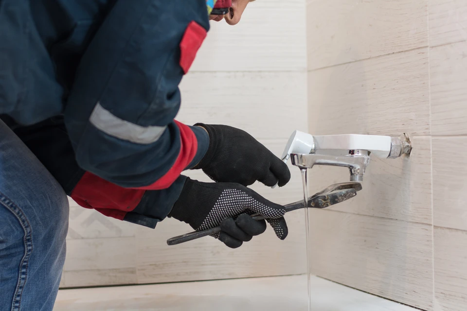 Plombier professionnel en combinaison bleue et gants noirs travaillant dans salle de bain carrelée blanche