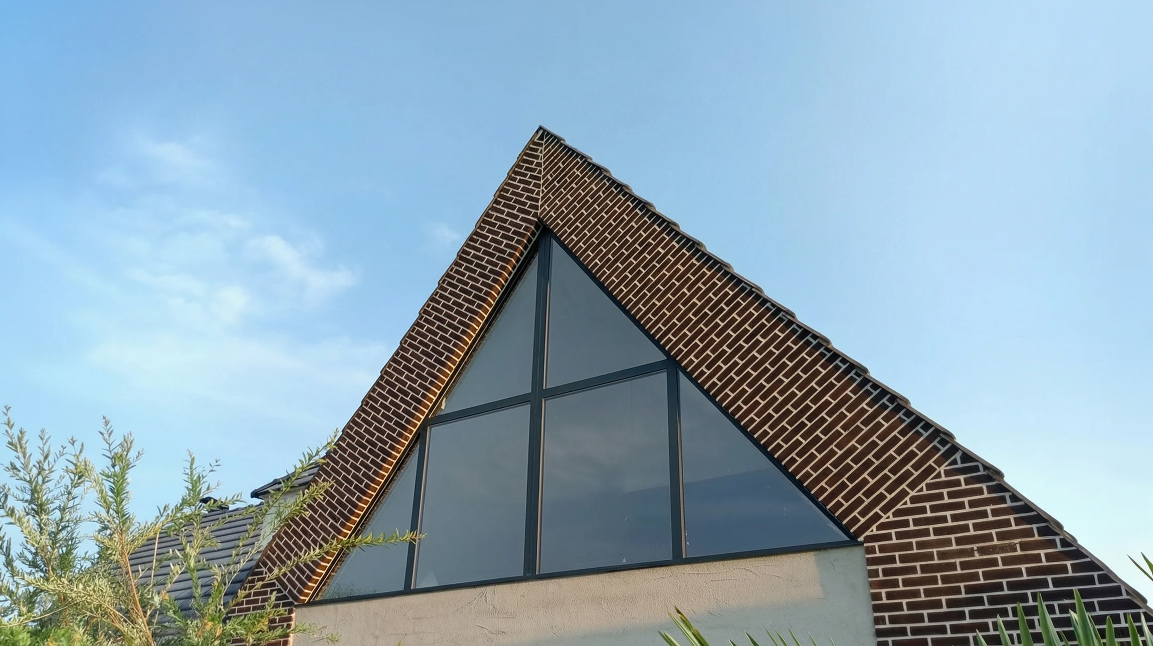 Fa&ccedil;ade ext&eacute;rieure d'une maison contemporaine avec mur en briques noires et grande verri&egrave;re triangulaire